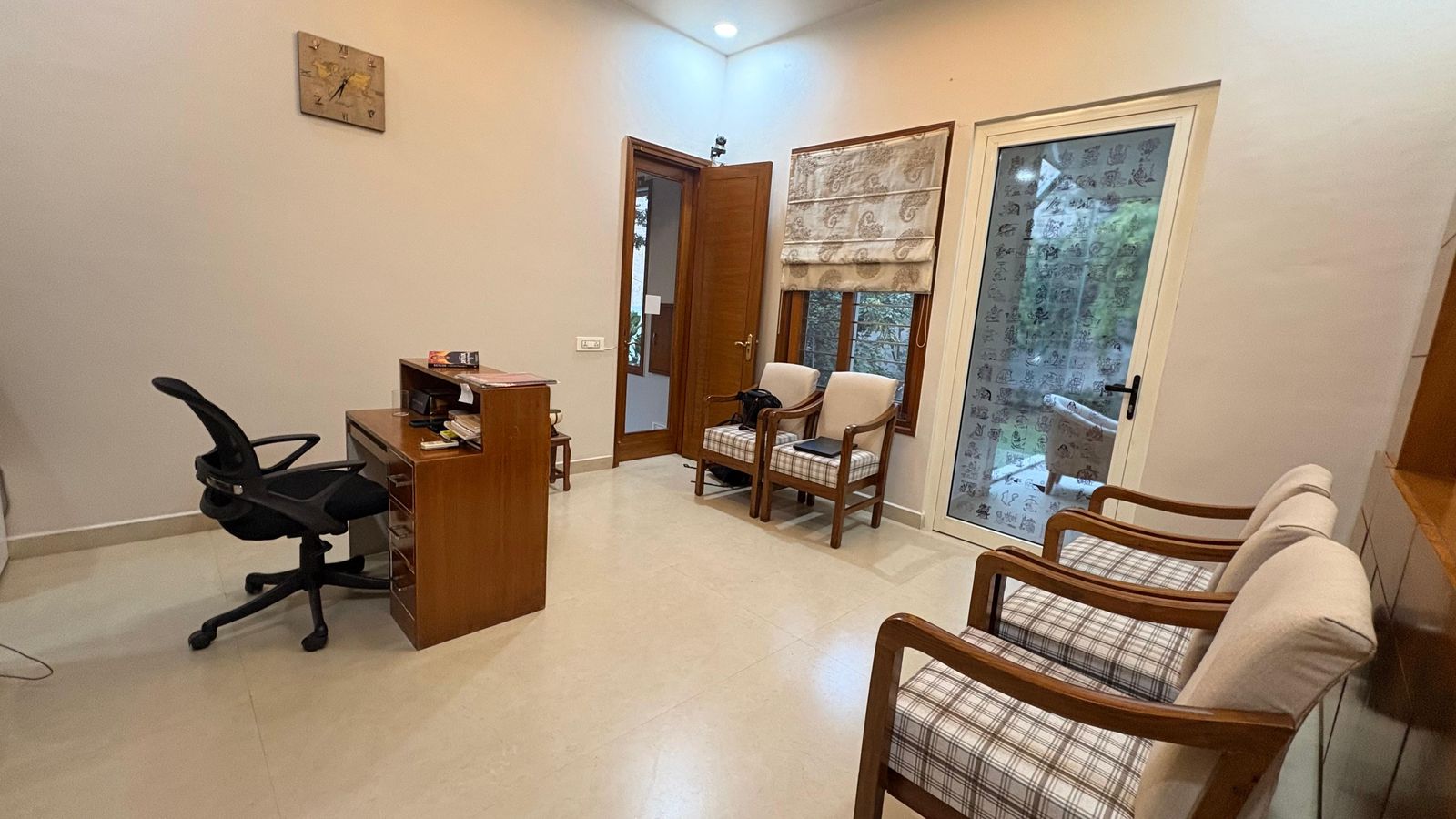 The consulting room from the door: the wooden desk and its chair, two upholstered chairs for visitors, and a framed yantra on the wall.