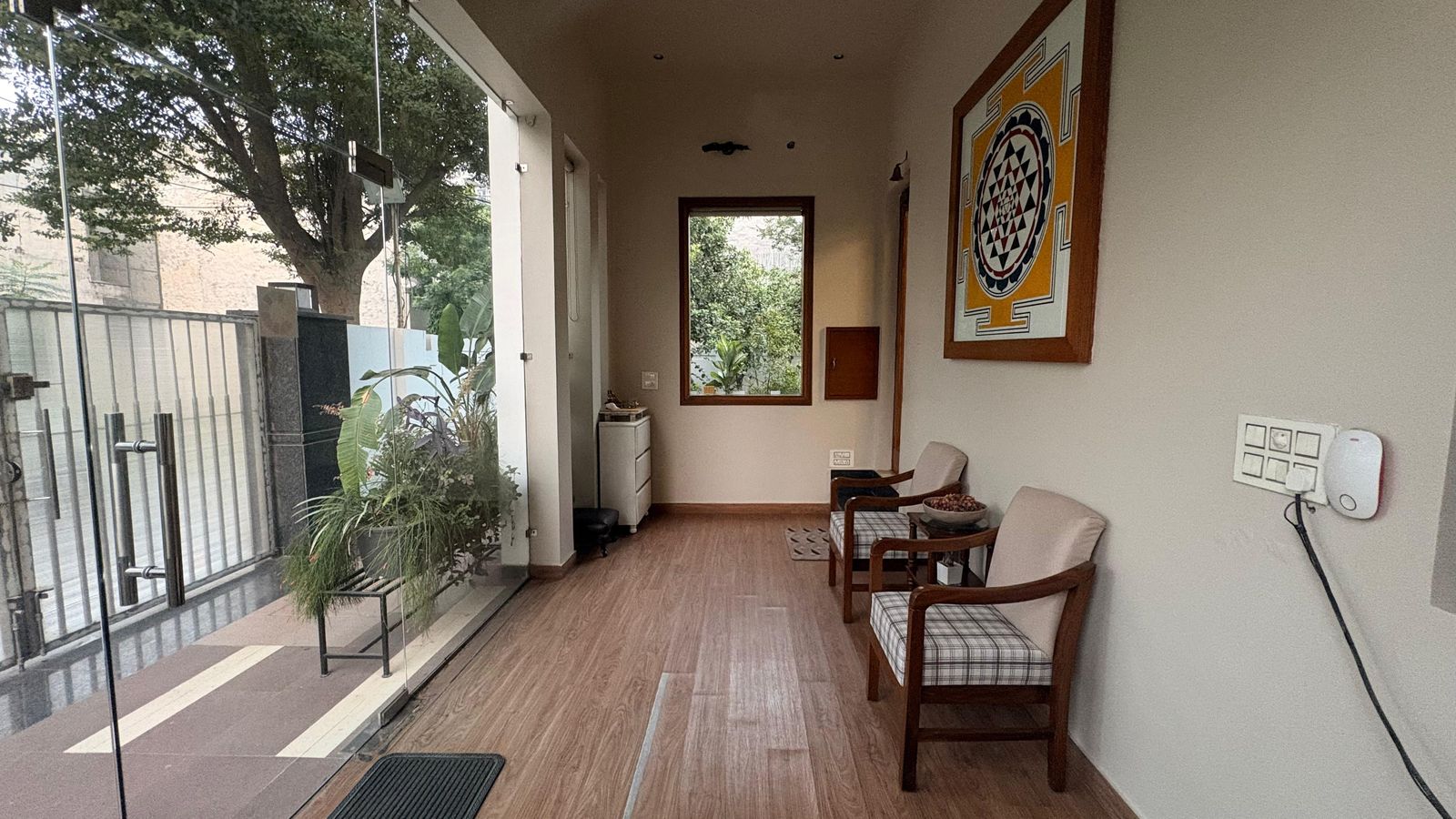 The entrance hall: a framed print on the wall, a chair, and the doorway open to the gate and the greenery beyond.