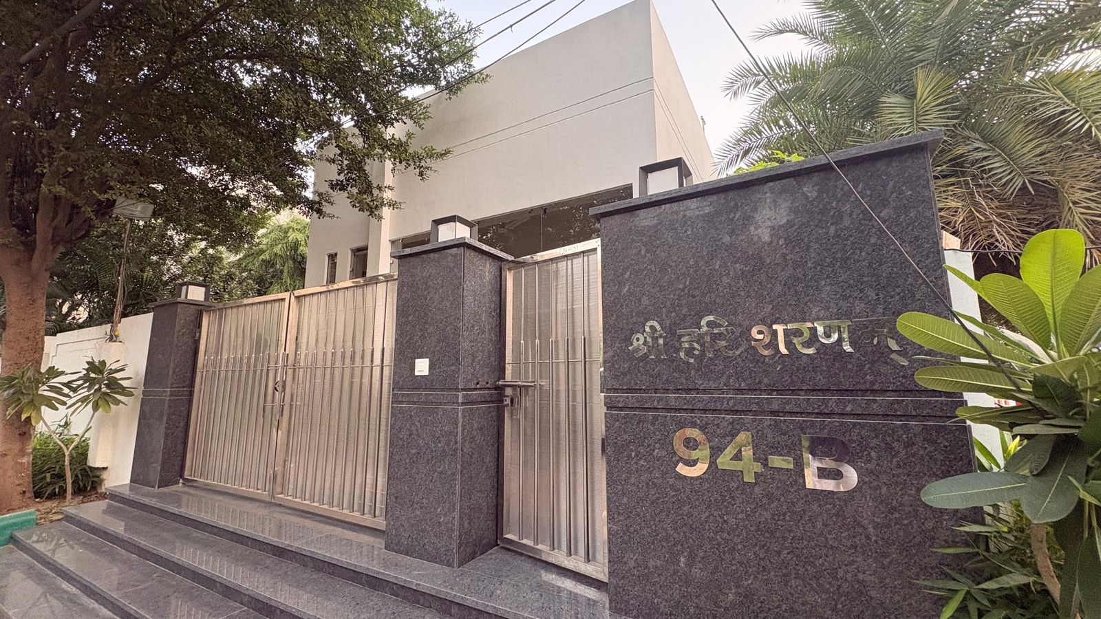The gate at 94-B, Kiran Vihar: a granite pillar with the house number, the Devanagari blessing above it, and the practice’s blue board beyond.
