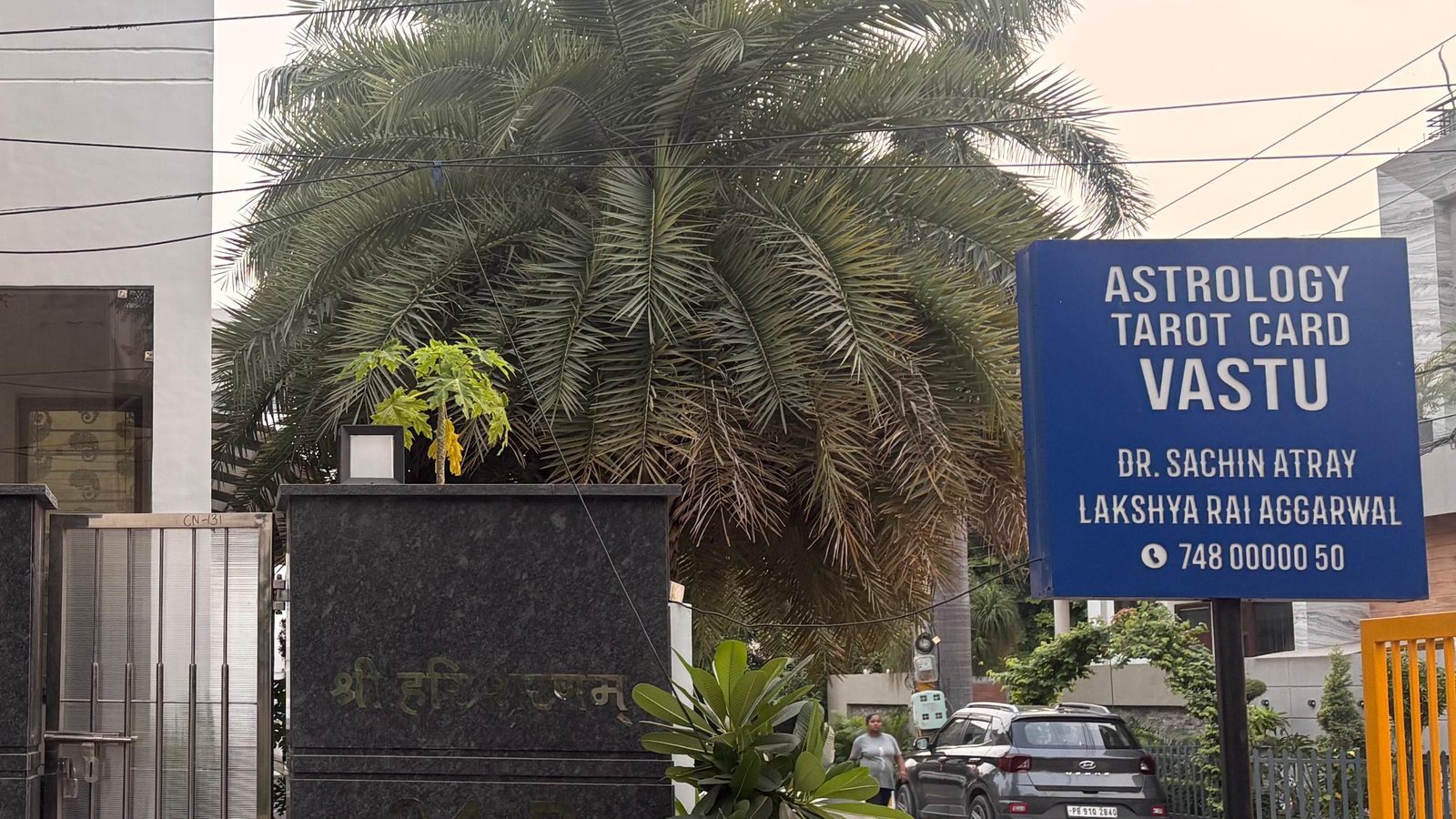 The practice’s board at the street gate, reading Astrology, Tarot Card, Vastu, with both readers’ names and the phone number.